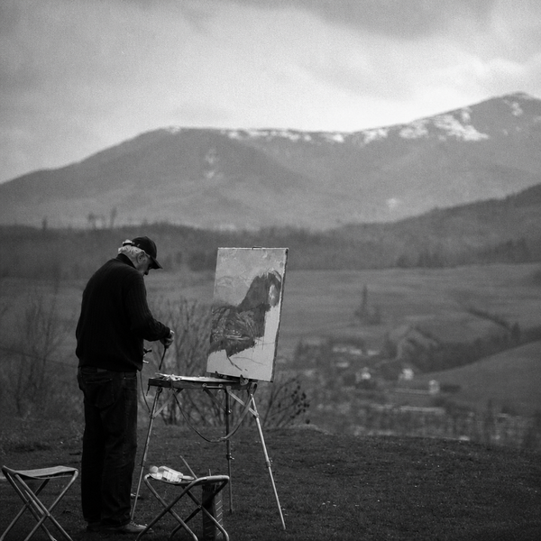 Black-and-white photograph of a painter working at a portable easel in an open field, mountains and a village behind him.