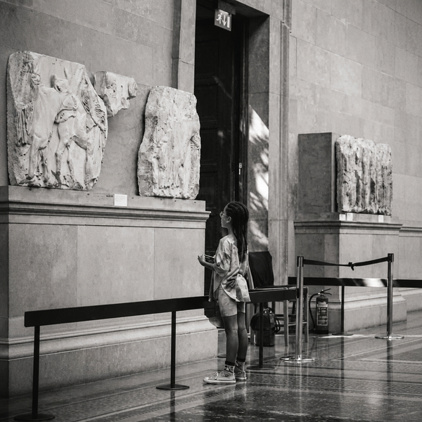A young girl in sneakers stands at a low barrier in a stone museum gallery, looking up toward a small label card beneath carved marble relief panels.