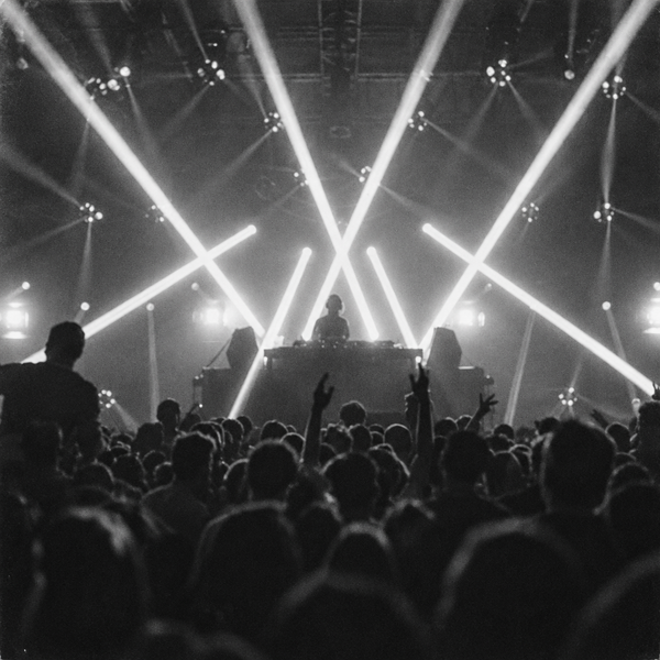 Black-and-white photo of a packed club crowd, some with hands raised, facing a DJ booth under crossing beams of stage light.