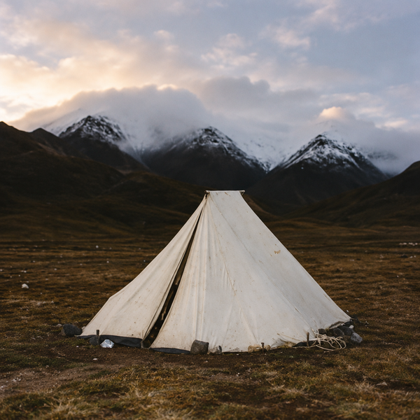 A weathered white canvas tent pitched alone on brown tundra, its edges weighed down with rocks, beneath snow-dusted mountains and low cloud.