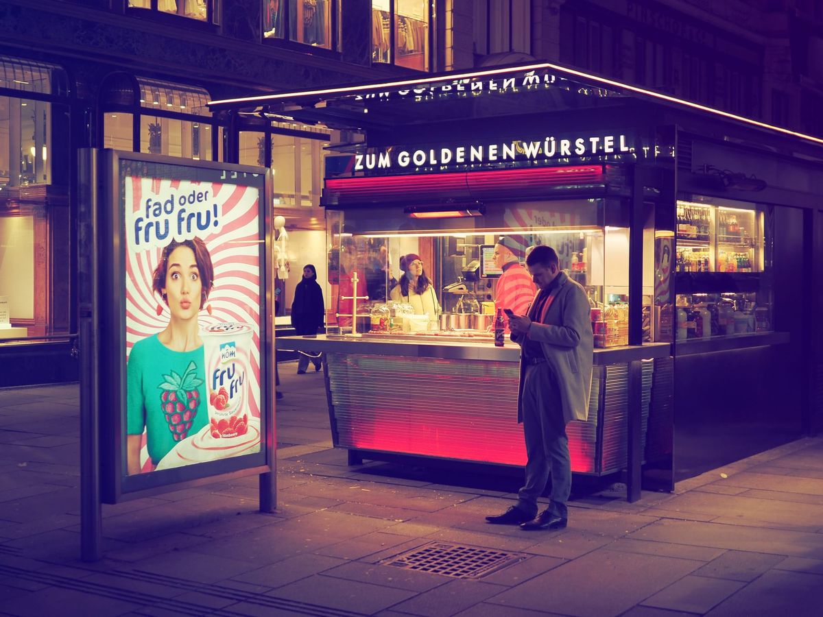 People standing at a street food stand at night in Vienna, with illuminated signage, warm light, and pedestrians nearby.
