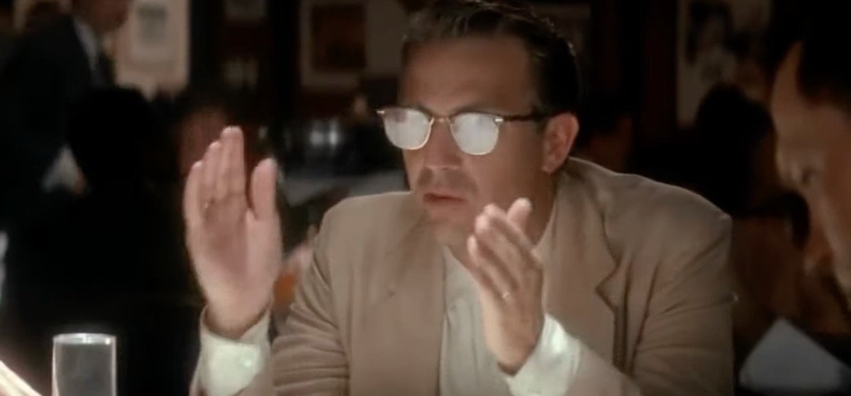 A still of Kevin Costner from JFK, in sunglasses, holding his hands up, explaining something