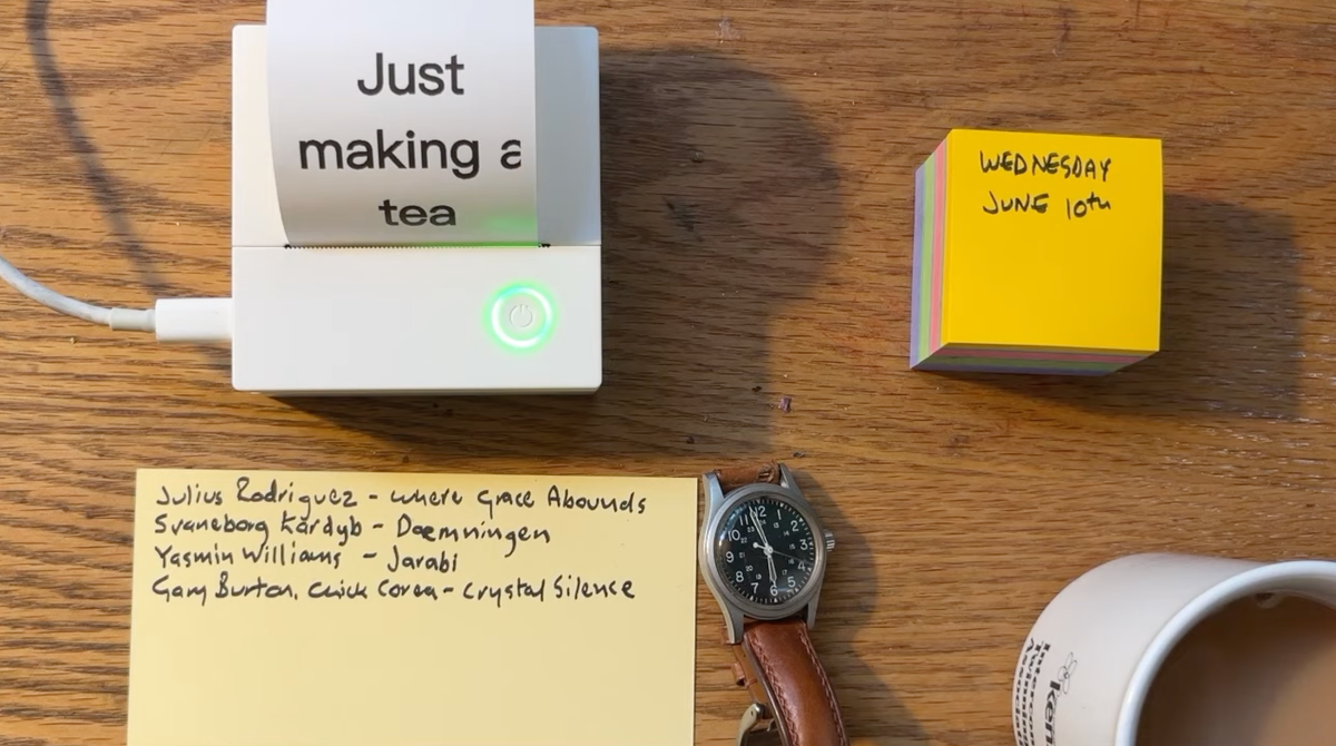 Looking down on a desktop with a watch, a cup of tea, a track list written on a card and a tiny printer with the text 'just making a tea'