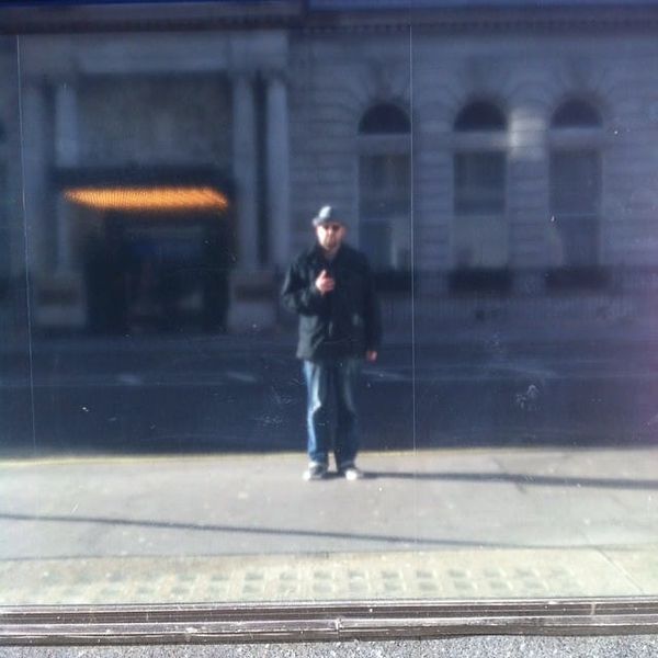 A man with a hat, black jacket and jeans, photographing himself in a blurry mirror, at some distance