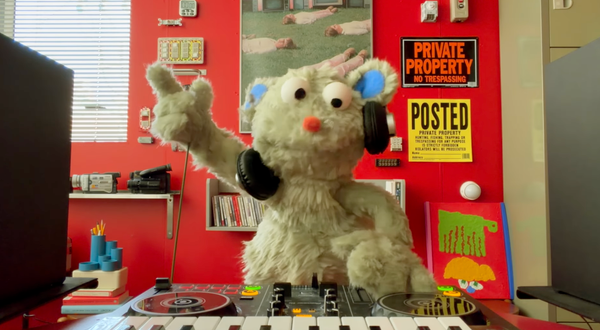 A small grey muppet like puppet behind some DJ decks