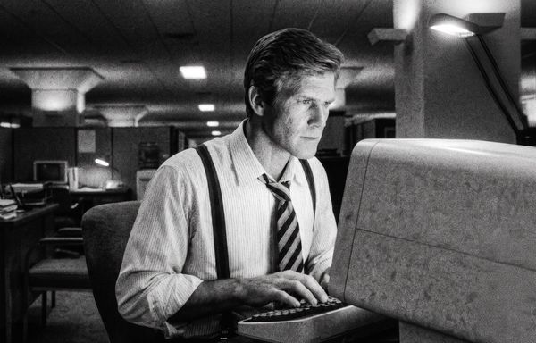 Grainy black and white photograph of a man in shirt, tie and suspenders at a CRT computer in an 1980s office, lit by a single desk lamp.