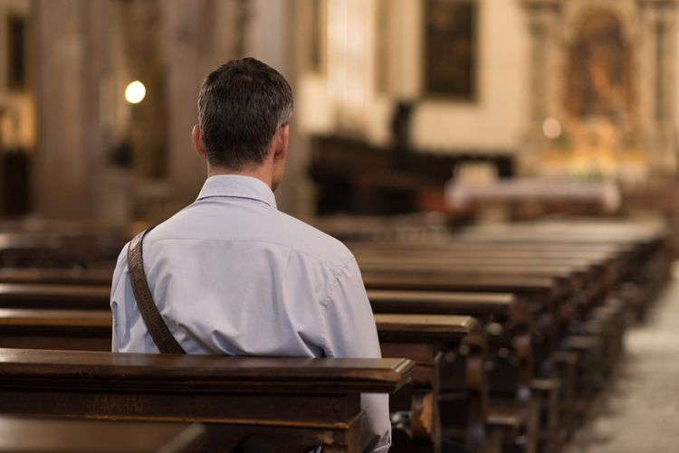 A man is sitting in a wooden pew at church facing the front and looking at the altar.