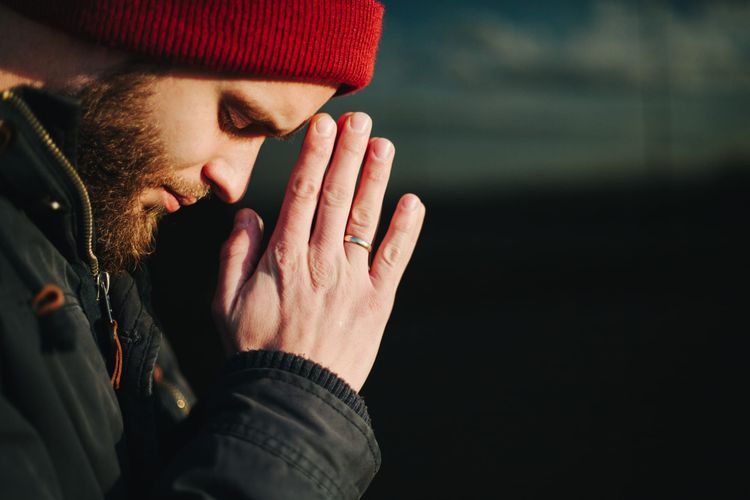 A man with a beard wearing a red knit cap has his hands folder near his face and his praying. 