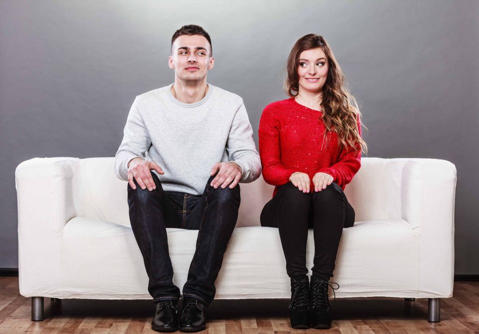 A shy man and woman looking awkwardly and sitting next to each other on a white sofa. 