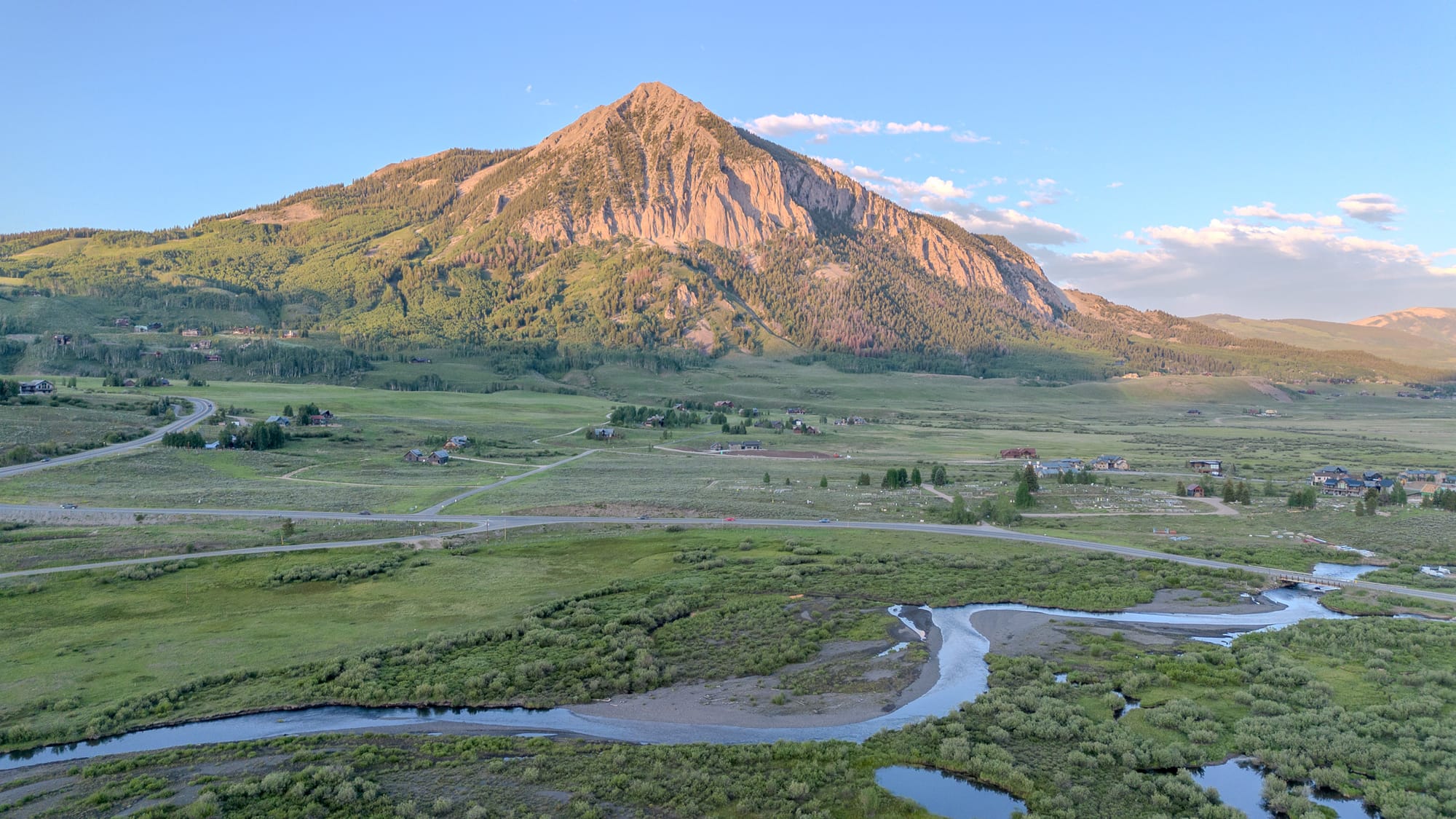 Photo of a mountain with a winding stream in the foreground. The sun is setting, and shadows fall on the mountains flanks, but the bulk of the mountain is sunlit in a peach color. 
