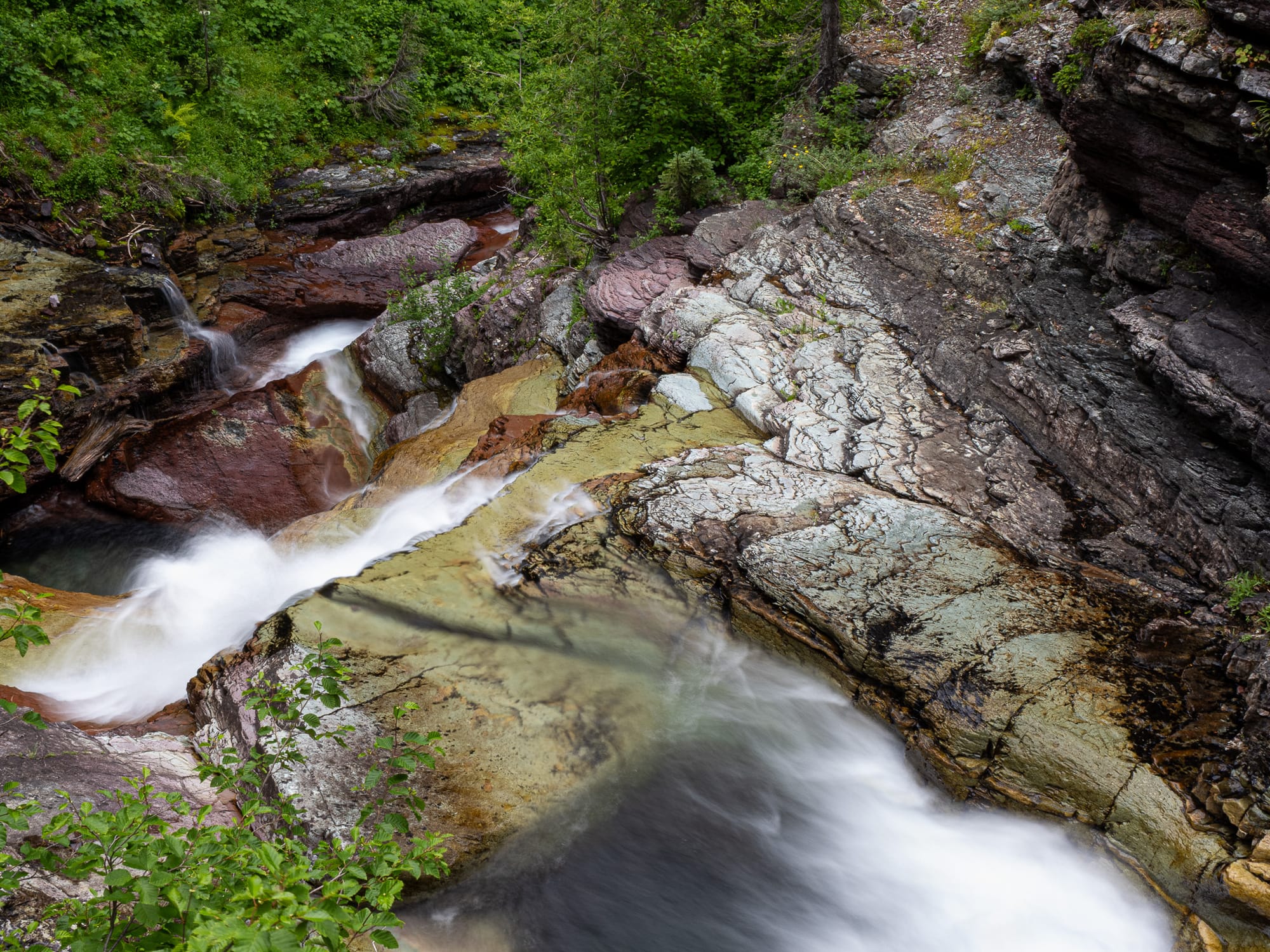 Water pours over a small waterfall viewed from above. There is green foliage and red stone. The water is blurred by a slow shutter. 