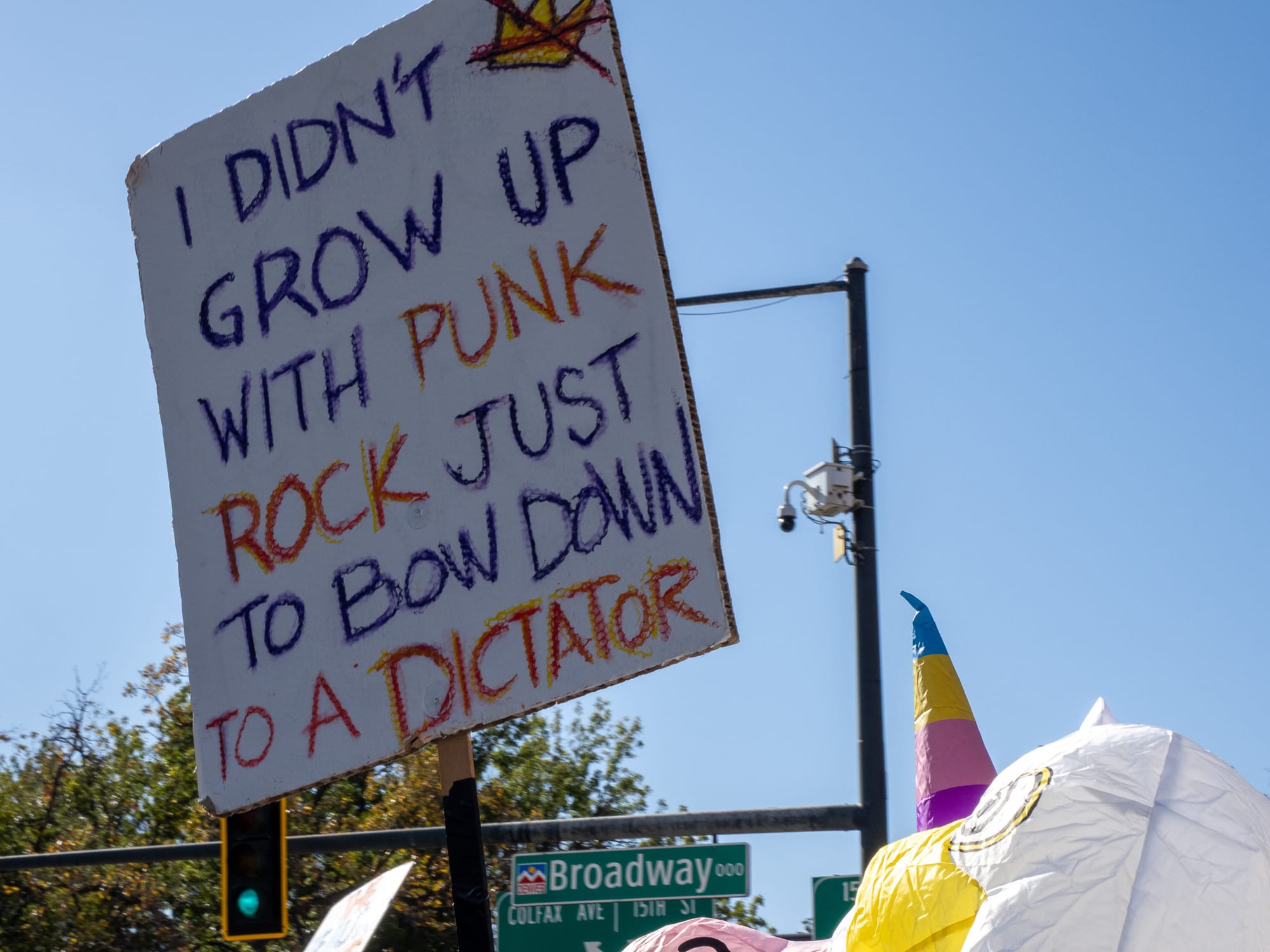 A protest sign that says “I didn’t grow up with punk rock just to bow down to a dictator” is held aloft against a blue sky.