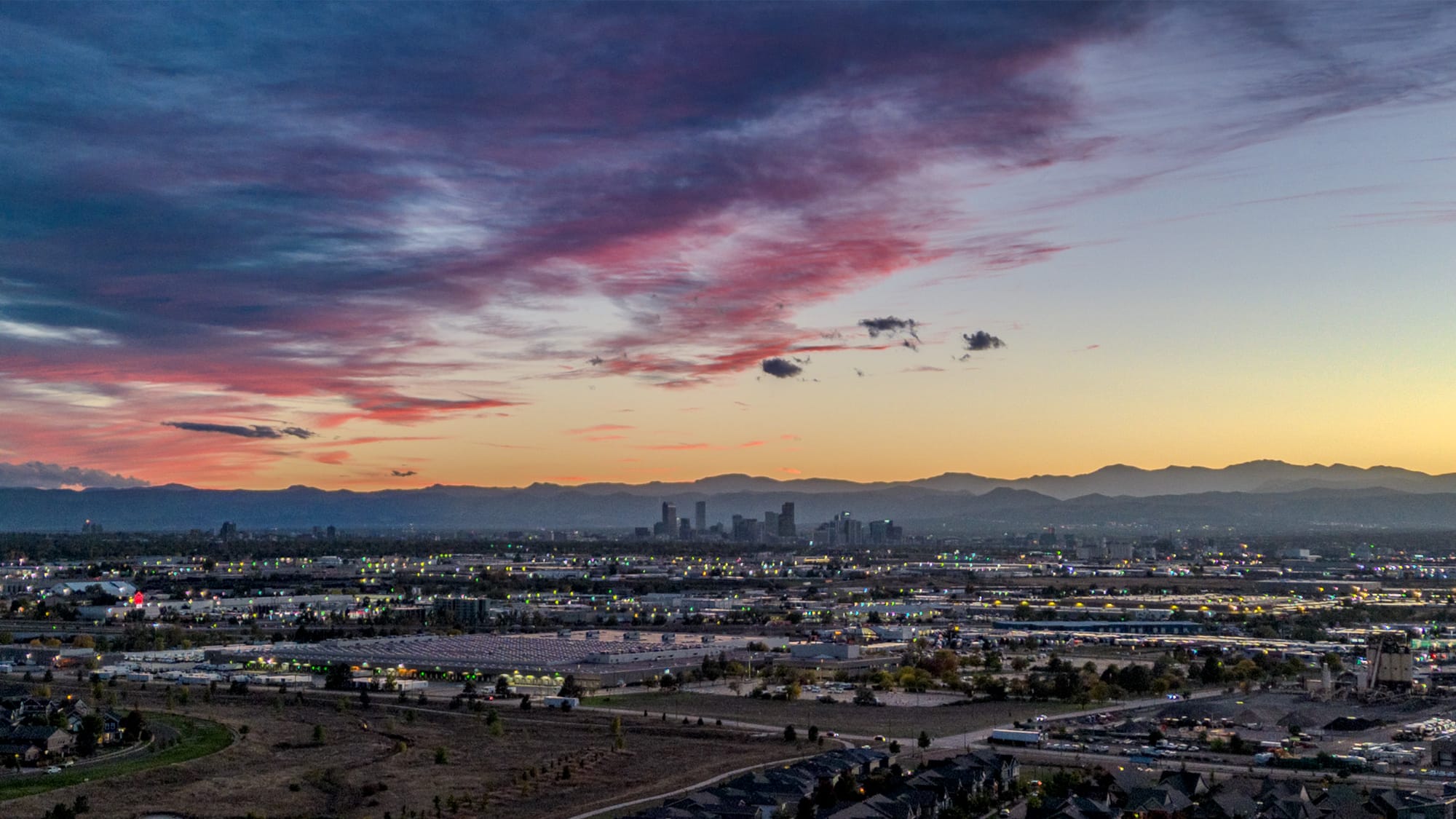 In the foreground, low buildings are visible with lights on, in the distance, a city skyline of tall buildings can be seen. Behind them, the rocky mountains are purple, with blue and orange skies, with a layer of red and purple clouds crossing from left to right. 