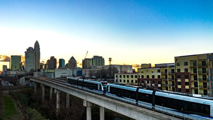 A light rail train crosses a concrete bridge in the foreground. In the background are apartments and a city skyline under blue and yellow skies as the sun sets.