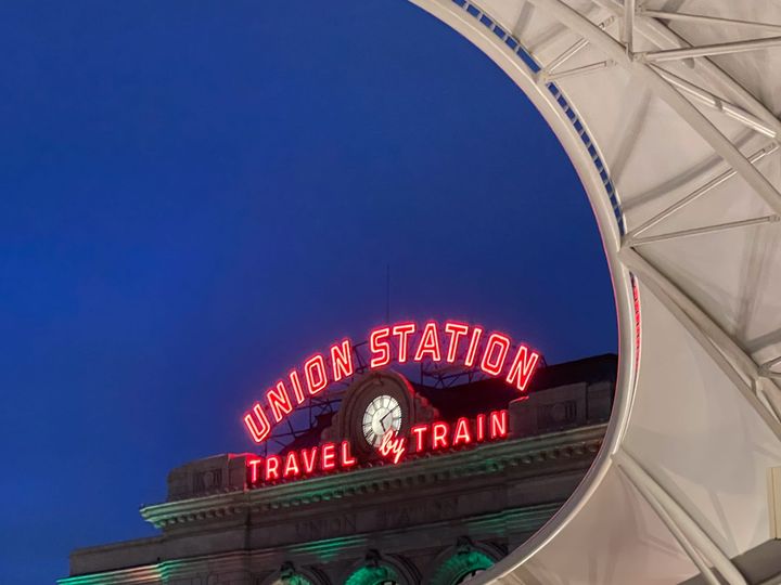 A red neon  sign under a purple nighttime sky says “Union Station Travel by Train”. A curving white awning frames the scene on the right. 