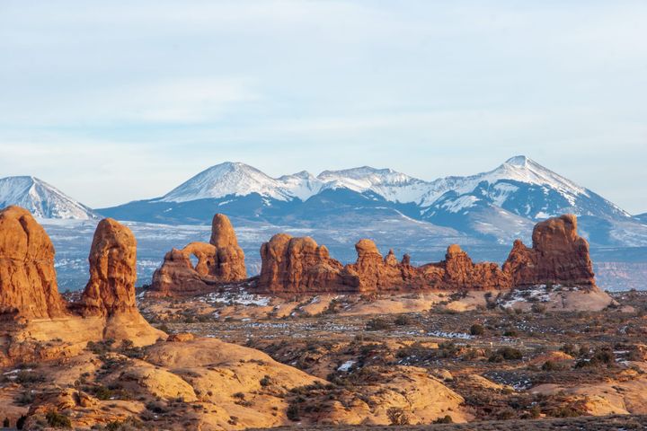 Red rock formations, including north window arch, dominate the fore and midground, while in the distance snow covered mountains rise above a treeline of dark colored trees
