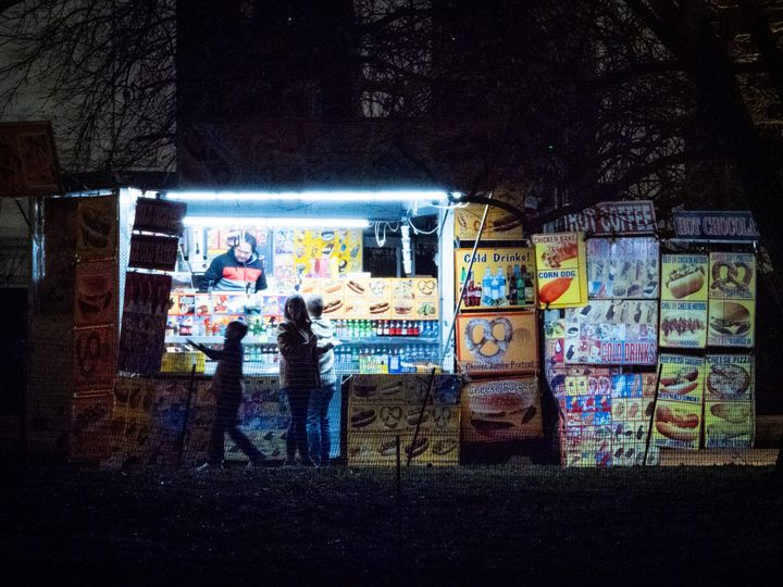 In a night scene, a food truck operator is illuminated by cool blue florescent lights while a family makes their selections. Signs on the right offer cold drinks, pretzels, and hot dogs. 