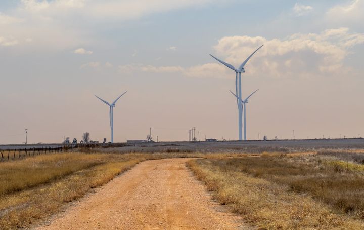 A dirt road in the foreground leads toward three large wind turbines under a hazy sky. 