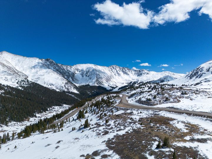A mountain scene under bright blue skies. In the foreground are large fields of snow sloping towards evergreen trees on the left.  