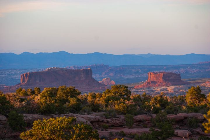 Red sandstone mesas are illuminated by the setting sun. Red rock terraces with green trees in the foreground. In the background, distant mountains are shaded blue by the setting sun. 