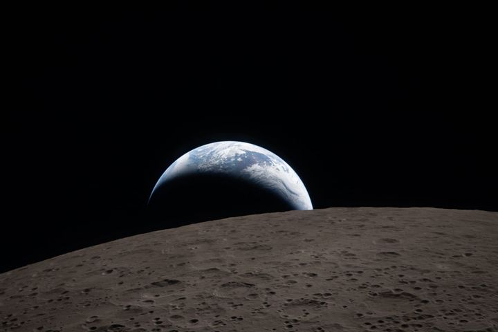 In the foreground, the heavily crated surface of the moon. In the background is earth, illuminated in a crescent of light. Blue oceans and white clouds are visible.