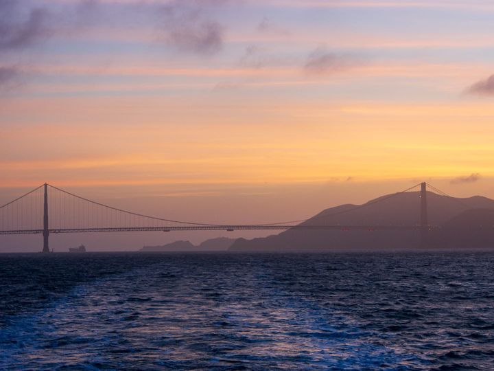 The Golden Gate Bridge is visible in the distance. There is dark blue water in the foreground and there are low clouds in purple, orange and blue from sunset. 