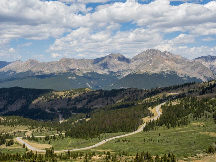 A mountain vista. In the foreground a road snakes up an alpine valley, while in the distance huge rocky mountains loom under while clouds in a blue sky.