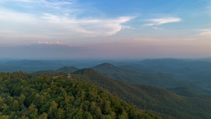 An aerial view of green, tree covered mountains under clouds and blue sky. 