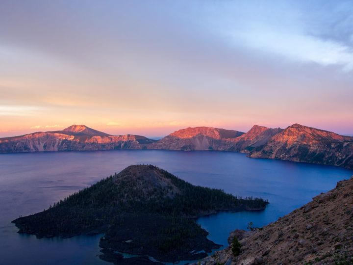 A mountainous island sits in a lake surrounded by mountains. The distant peaks are lit with orange and red alpenglow. The lake water is a deep blue.