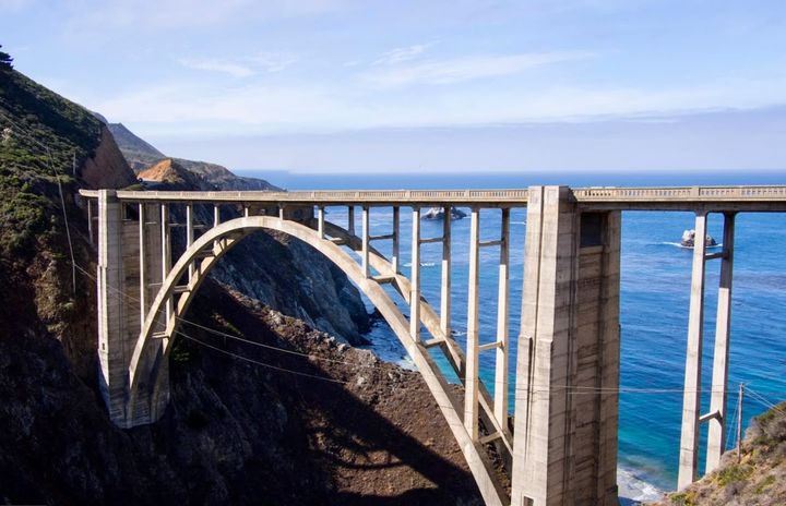 A concrete arch bridge spans a deep canyon. Behind the bridge is the Pacific Ocean. The ocean is deep blue. 