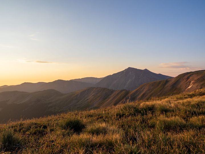 A mountain scene at sunrise. In the foreground are golden grasses. A ridge line extends into the distance. Behind the ridge rises another taller mountain
