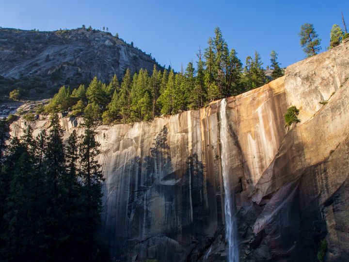 A thin stream of water pours over the lip of a large cliff. Evergreen trees and the top of the cliff catch the evening light. 