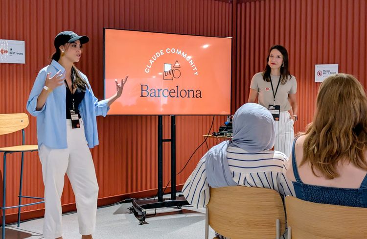 Two female speakers stand in front of a television that says Claude Community Barcelona, speaking to a crowd of workshop participants.