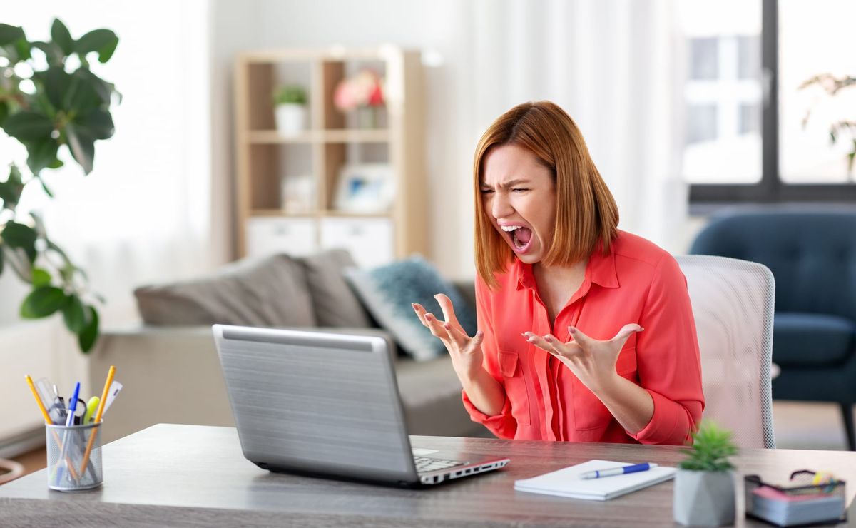 A red headed woman in a pink blouse sitting in an office chair at a gray desk with office supplies on it and a gray laptop in front of her. She's screaming and mad.