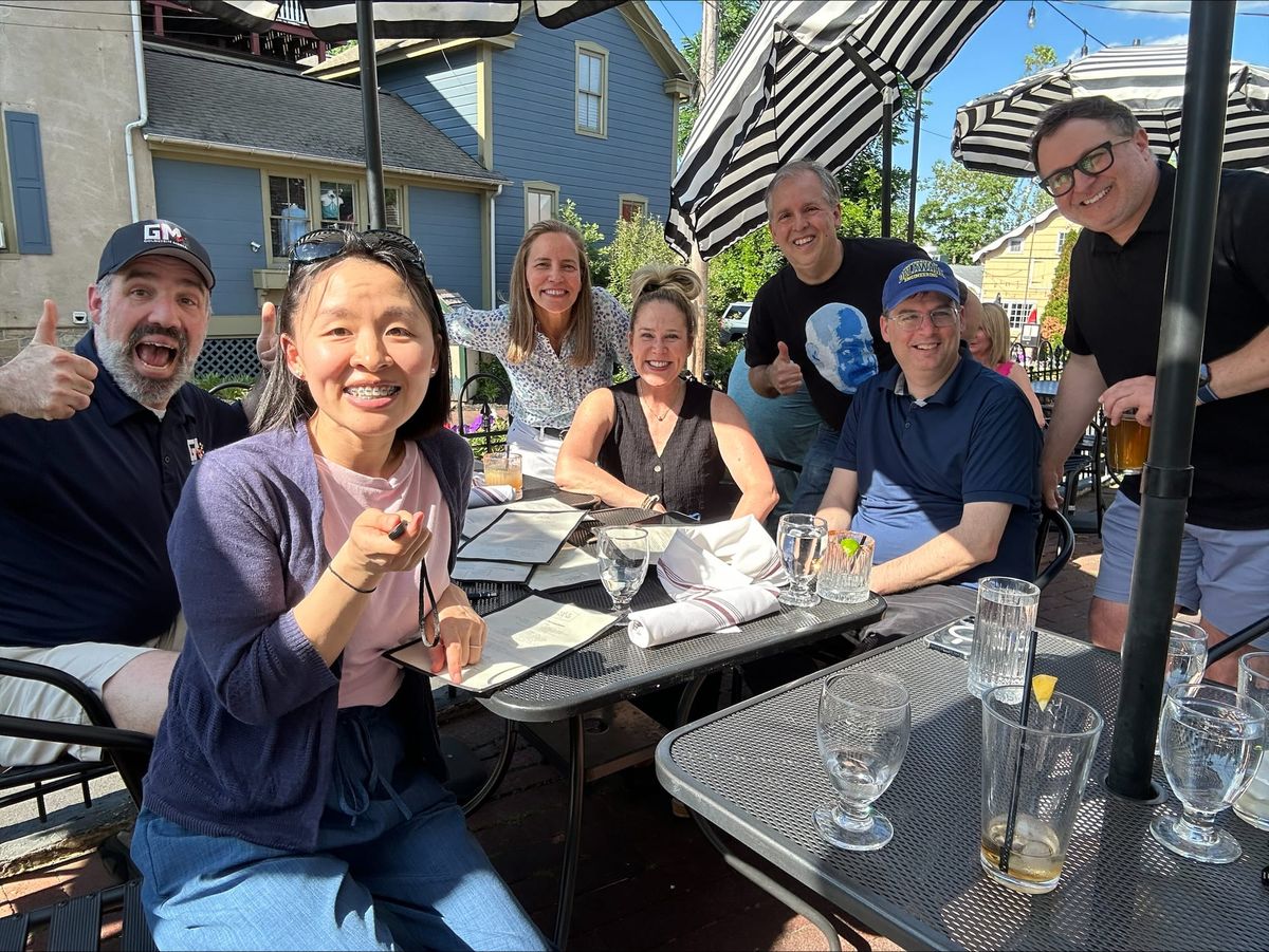 A group of people sitting at a table taking a group shot. All smiling for the photo. Beverages and menus are on the metal mesh tables.