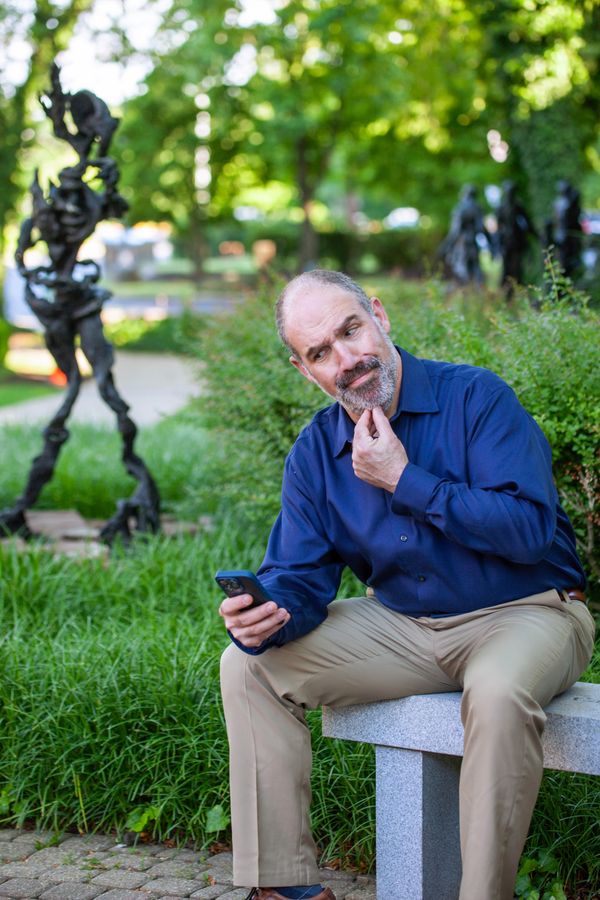 Seth looking at his phone with suspicion. He's sitting outside on a stone bench with green foliage around him and cobble stones. There are sculptures behind him.