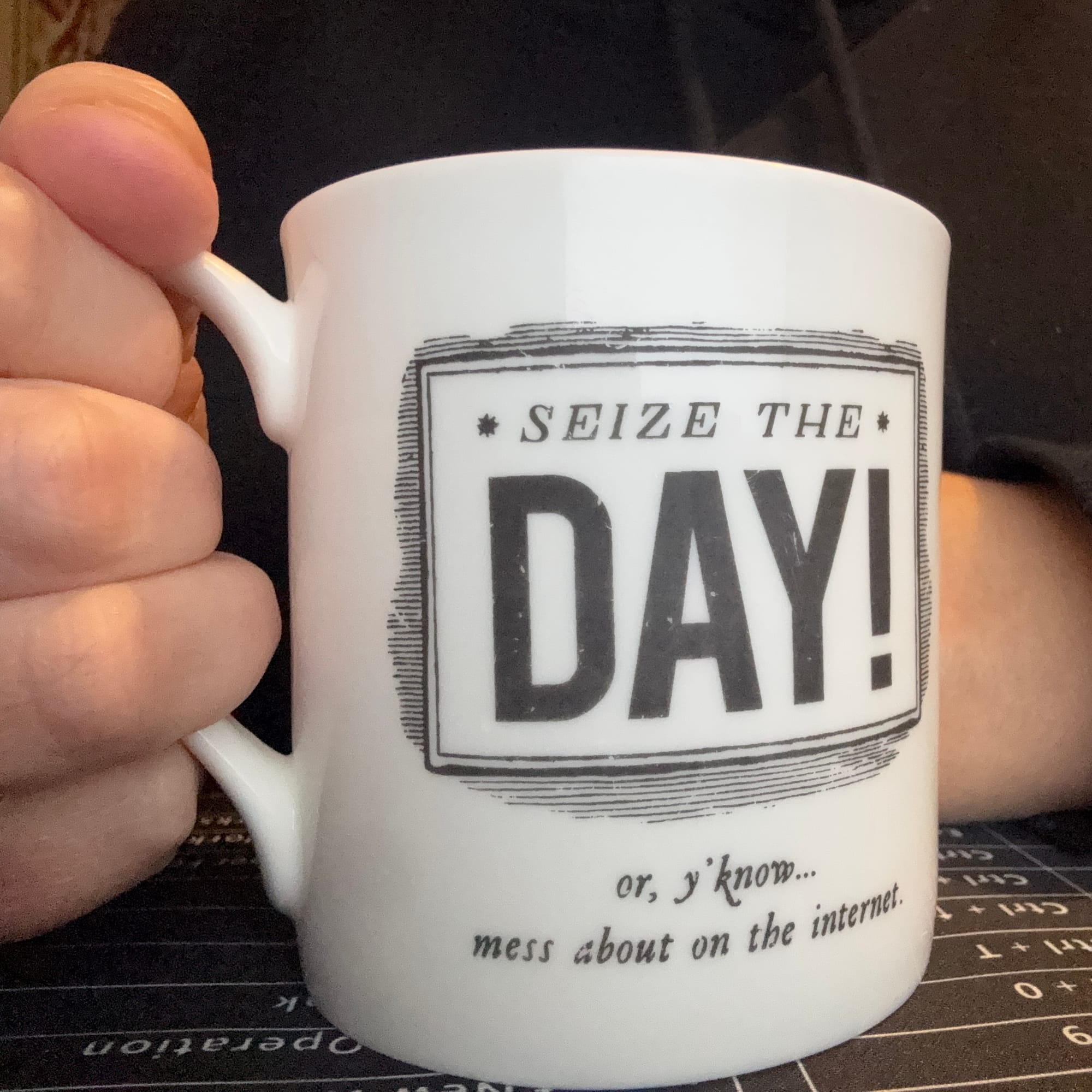 A picture of Gayle (well, her black hoodie and hands, you can't see her face) holding her (actual) cup of tea this morning, on her keyboard shortcut desk mat. Her mug says "Seize the day! or, y'know... mess about on the internet"