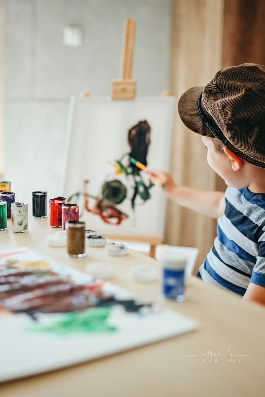young boy sitting at table with paints, an easel, and a paintbrush in hand