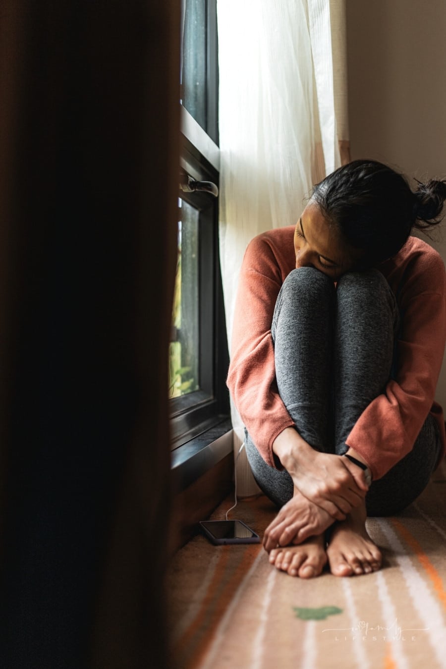 woman sitting by window hugging her knees to her chest