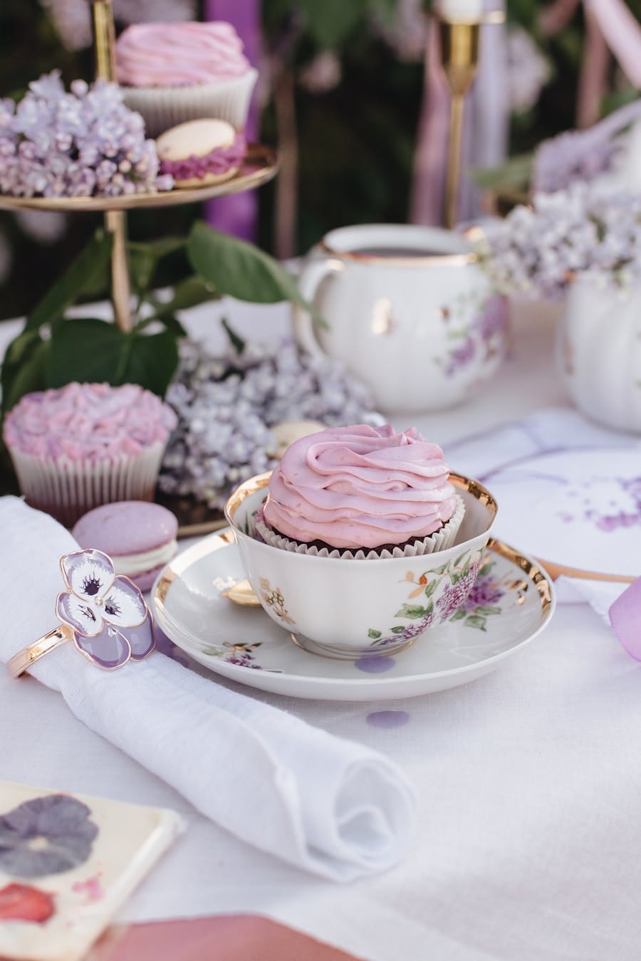 A delicate pink cupcake in an ornate teacup on a beautifully set table with a floral theme.