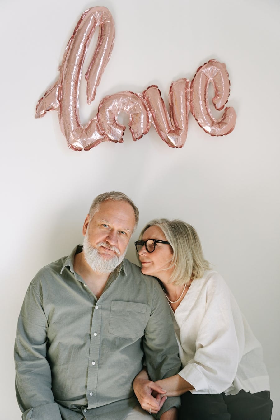 A loving elderly couple holding hands under a 'love' balloon indoors.