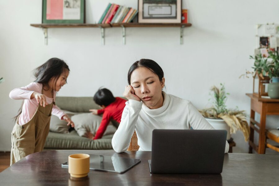 A tired mother working remotely on her laptop while children play in the background.