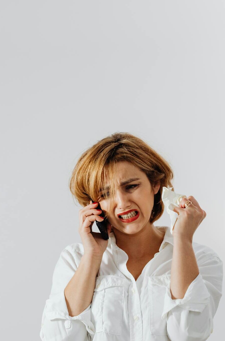 A woman expressing frustration during a phone call, wearing white long sleeves against a plain background.