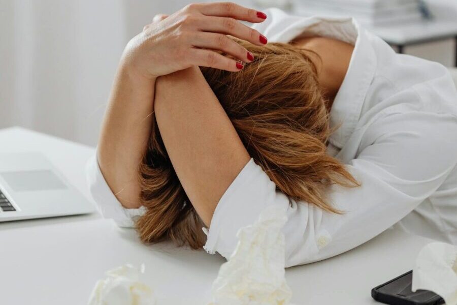 A woman in distress sits at a desk with head in hands, surrounded by tissue papers.