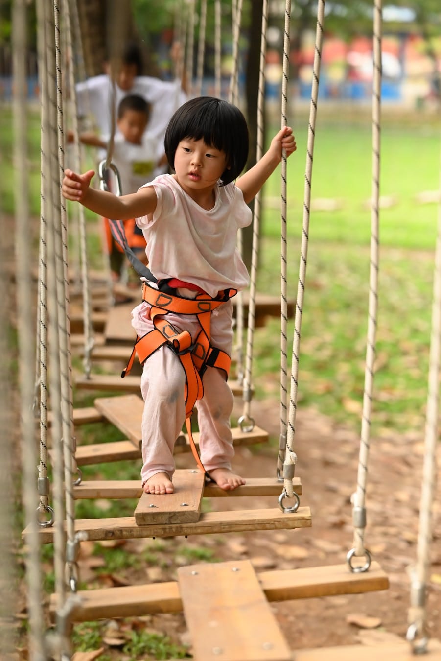 A young girl carefully crossing a rope bridge at an outdoor playground, wearing a safety harness.