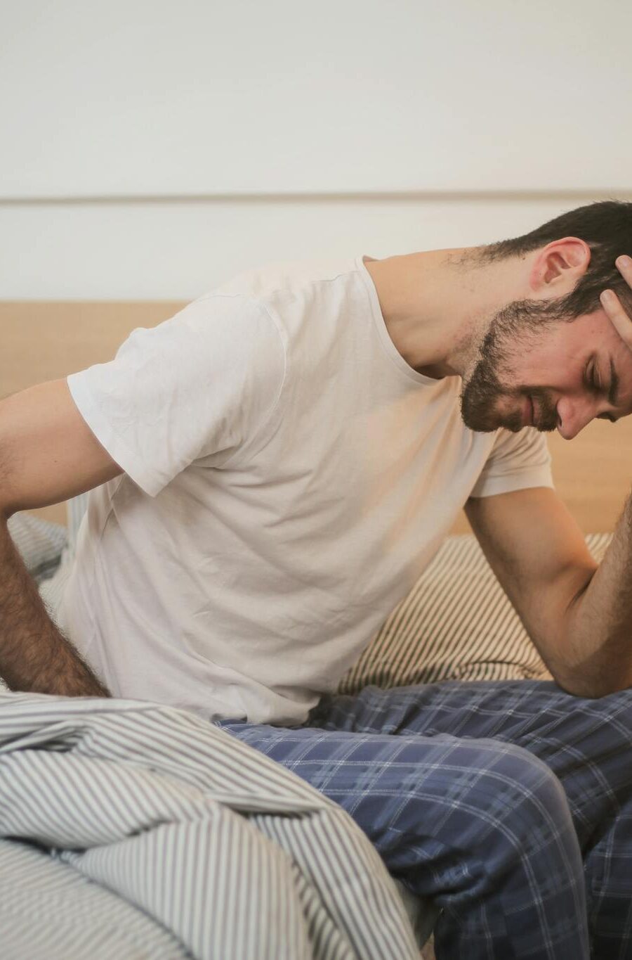 A young man in pajamas holding his head, sitting on a bed, appears to be experiencing a headache.