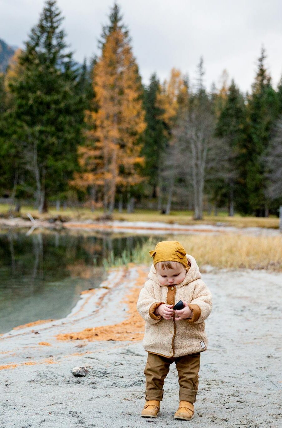 Adorable toddler in a warm jacket playing by a serene lakeside in autumn scenery.