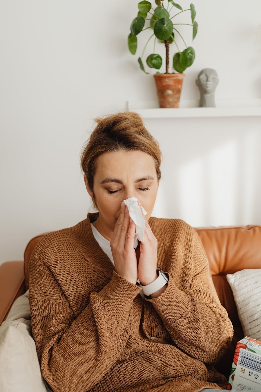 Adult woman in a sweater blows her nose while sitting on a leather sofa in a cozy living room setting.