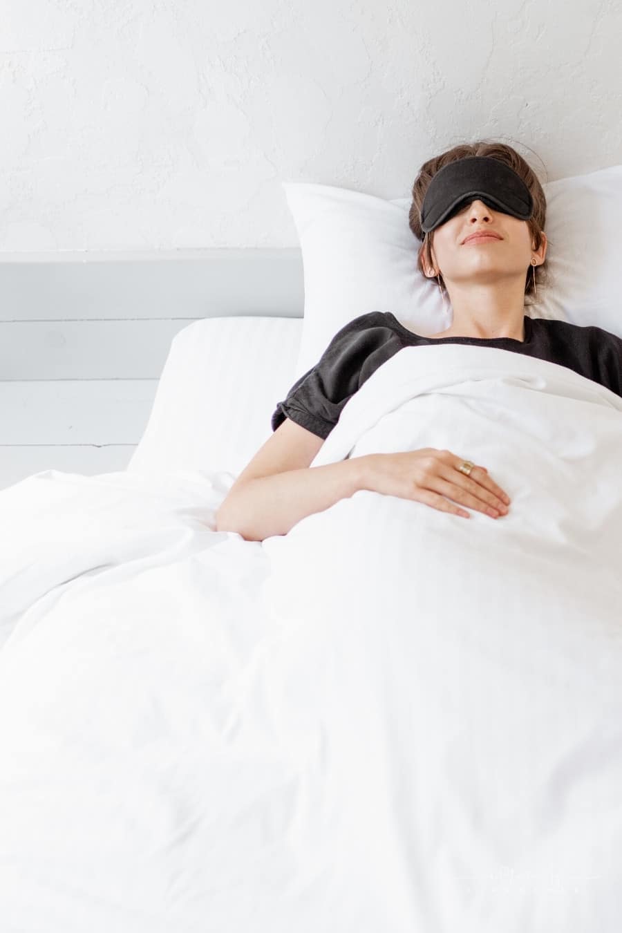 woman lying with a sleeping mask in bed under white comforter blanket