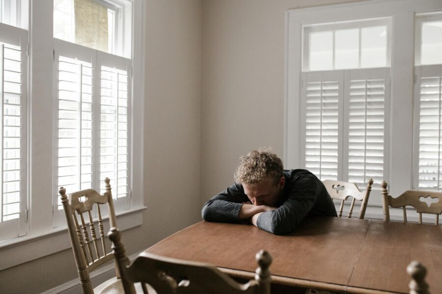 An adult man with emotions of sadness and anxiety sitting alone at a wooden table near windows.